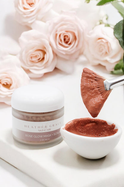 Open jar of Heather Grace Rose Glow Mask + Scrub alongside a brush dipped in the product, set against a backdrop of soft pink roses, depicting a ready-to-use skincare treatment.