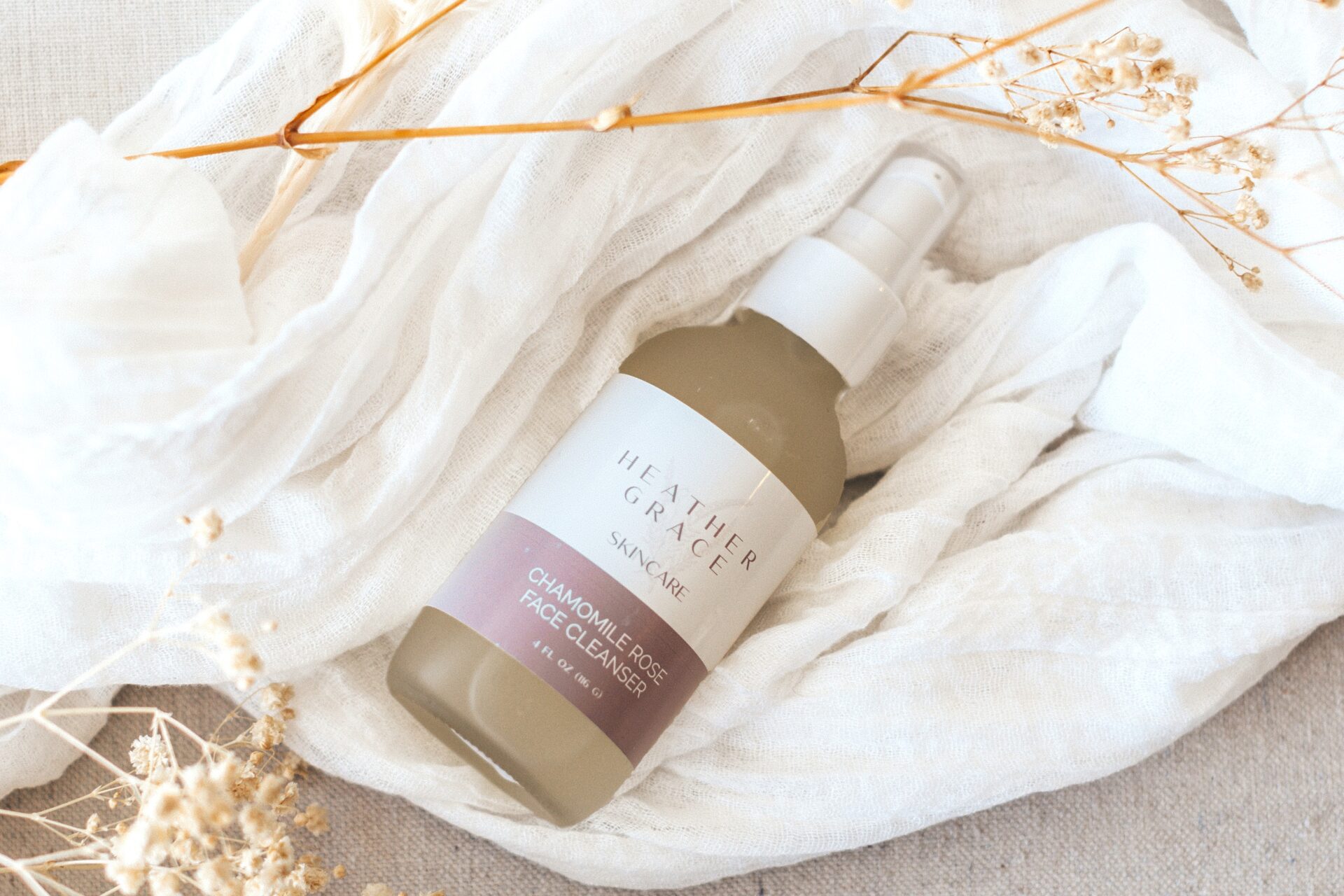 A bottle of Heather Grace Chamomile Rose Face Cleanser sits atop a crinkled white fabric, accompanied by delicate dried botanicals. The cleanser's label is elegant and clear, with a serene and natural aesthetic in the composition.