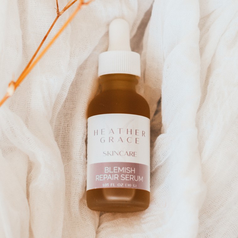 A dropper bottle of Heather Grace Skincare Blemish Repair Serum rests on a soft, white gauzy fabric, accompanied by delicate dried branches, creating a serene and natural setting that evokes a sense of gentle care for the skin