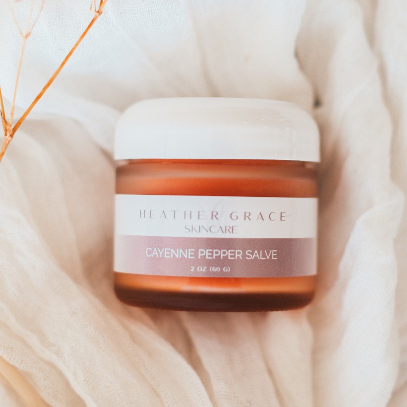 A jar of Heather Grace Cayenne Pepper Salve is nestled on a soft, white, textured cloth with faint dried plant stems in the background, highlighting the product's natural and soothing essence.