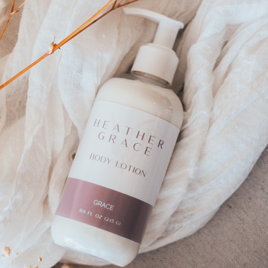 A pump bottle of Heather Grace Body Lotion titled 'GRACE' is elegantly placed on a crumpled white gauzy fabric, with warm-toned dried twigs adding a natural contrast to the soothing aesthetic of the product presentation