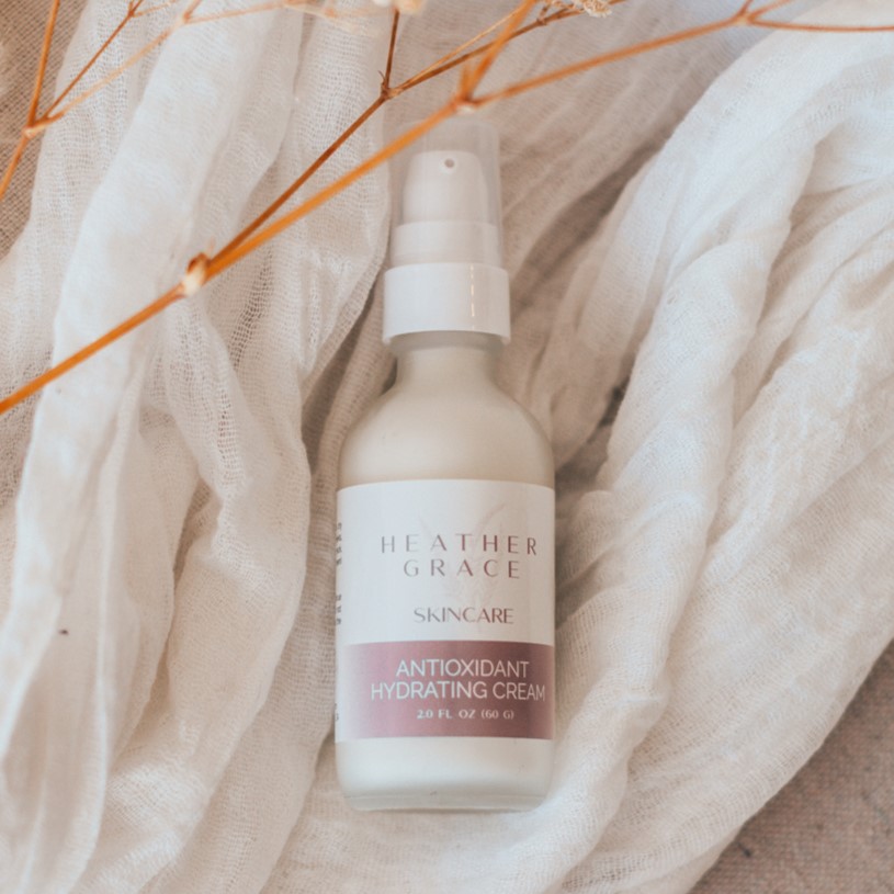 A frosted glass bottle of Heather Grace Antioxidant Hydrating Cream is placed on a delicate white gauzy fabric, with sprigs of dried plants adding a touch of earthy warmth to the soft, tranquil scene."
