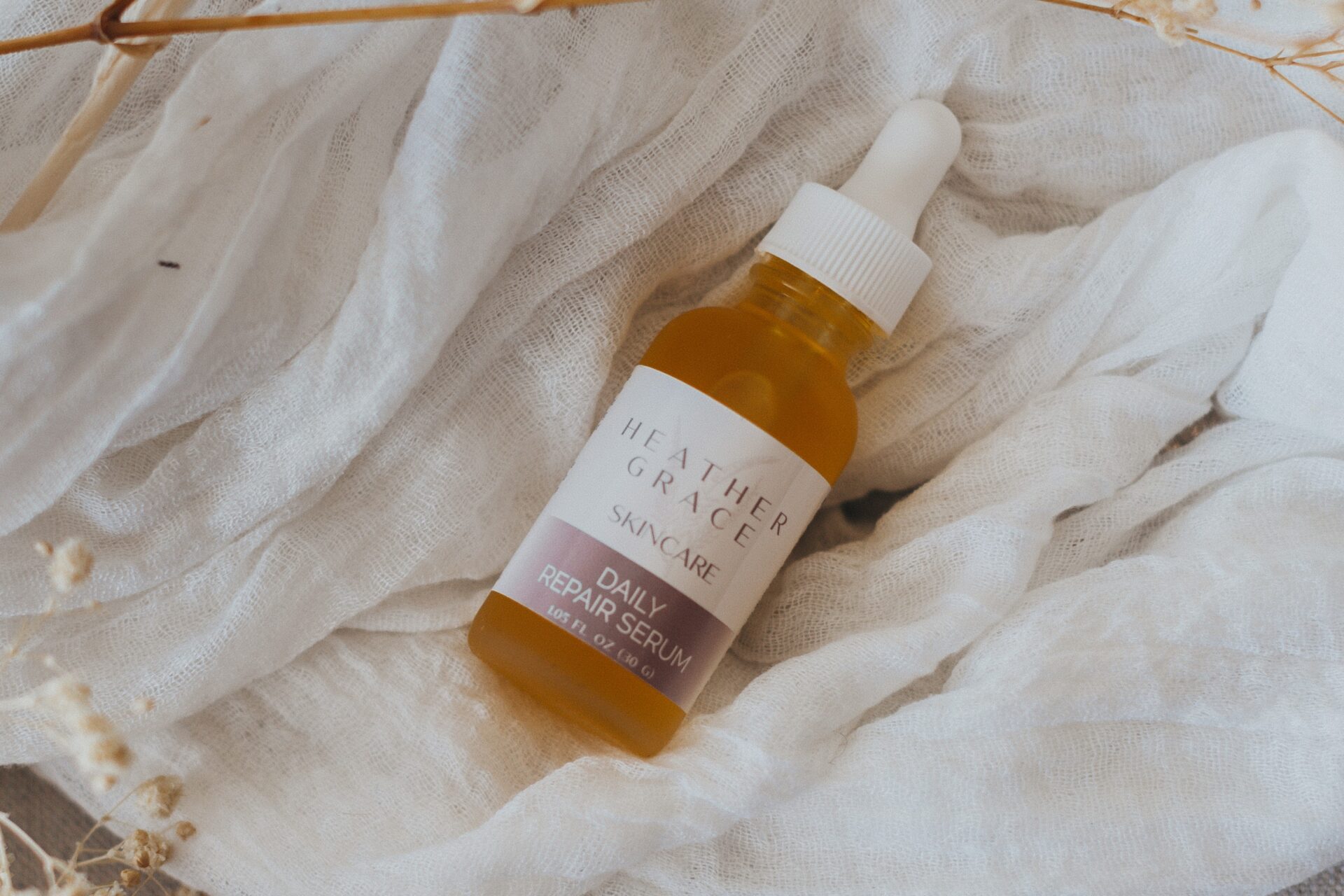 A bottle of Heather Grace Skincare Daily Repair Serum is nestled gently among folds of a soft, white gauzy fabric. Delicate dried baby's breath and wispy pampas grass in muted autumnal hues add a natural and serene touch to the composition, evoking a sense of gentle care and organic beauty. The scene suggests a peaceful, calming atmosphere, reminiscent of a quiet moment of self-care and rejuvenation.