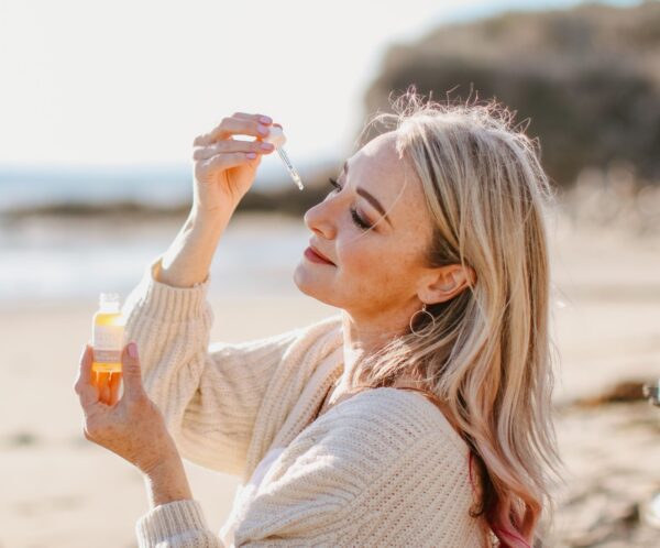 A woman with long blonde hair with pink ends is holding a small dropper bottle of amber-colored liquid up to the light, closely examining the contents. She's outdoors on a sunny beach, wearing a cream knitted cardigan over a white dress. Her expression is one of serene contentment, with her eyes gently closed and a soft smile, as she's surrounded by a tranquil coastal landscape
