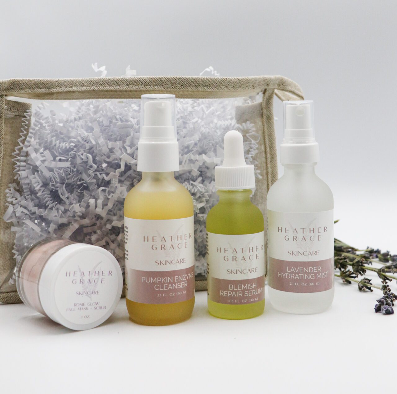 A collection of Heather Grace Skincare products displayed elegantly against a white backdrop. The set includes a 'Pumpkin Enzyme Cleanser', a 'Blemish Repair Serum', and a 'Lavender Hydrating Mist'. Beside them is a white jar labeled 'Rose Glow Face Mask & Scrub'. The products are accompanied by a rustic transparent pouch filled with white paper shreds and fresh sprigs of lavender scattered to the side.