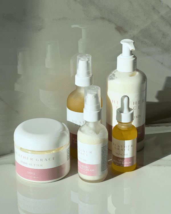 An elegant array of Heather Grace Skincare products displayed against a shadowy background with natural light casting a glow. The collection includes a large jar of body butter, a spray bottle, a pump bottle, and a dropper bottle, all with chic white and pink labels.