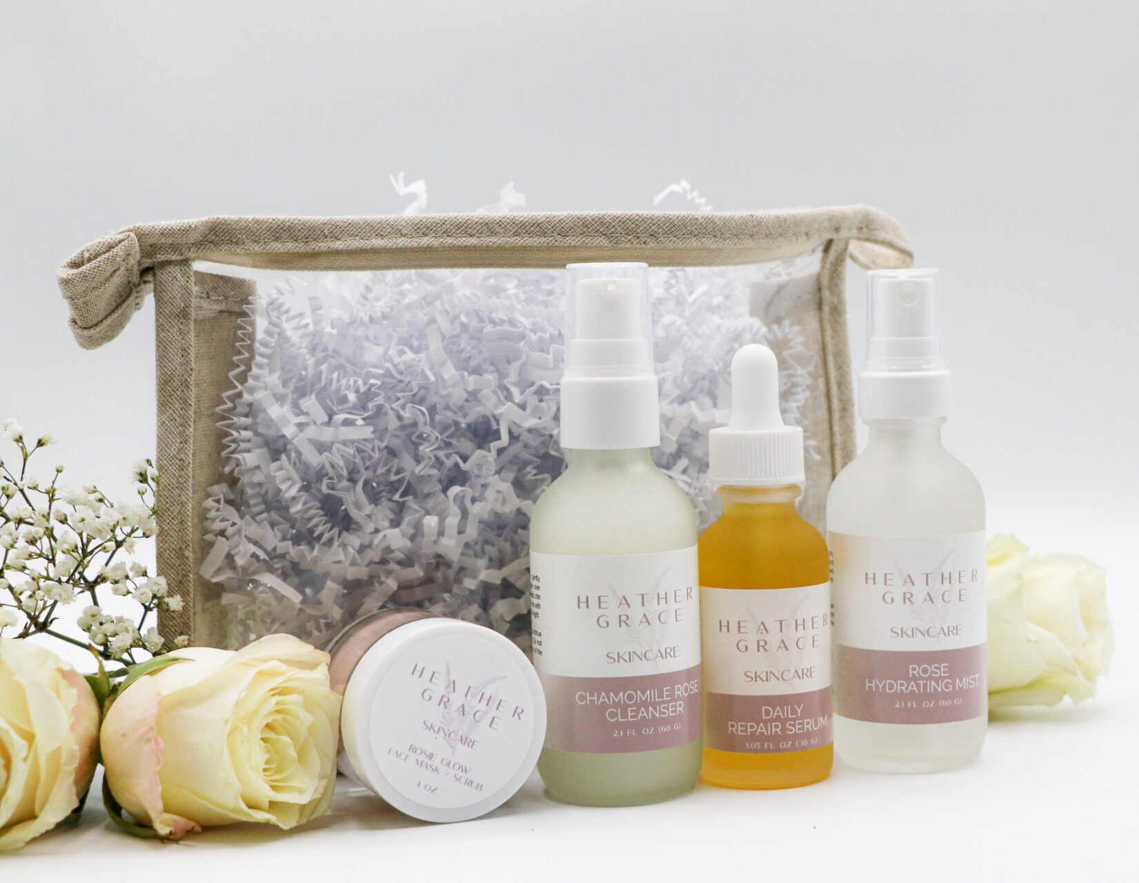 A skincare set from "Heather Grace Skincare" displayed against a white background. The set includes a "Chamomile Rose Cleanser", a "Daily Repair Serum", and a "Rose Hydrating Mist". Adjacent to the products is a white jar labeled "Rose Glow Facial Mask + Scrub". The products are arranged next to a transparent pouch filled with white paper shreds, fresh white baby's breath flowers, and two soft yellow roses. Is this conversation helpful so far?