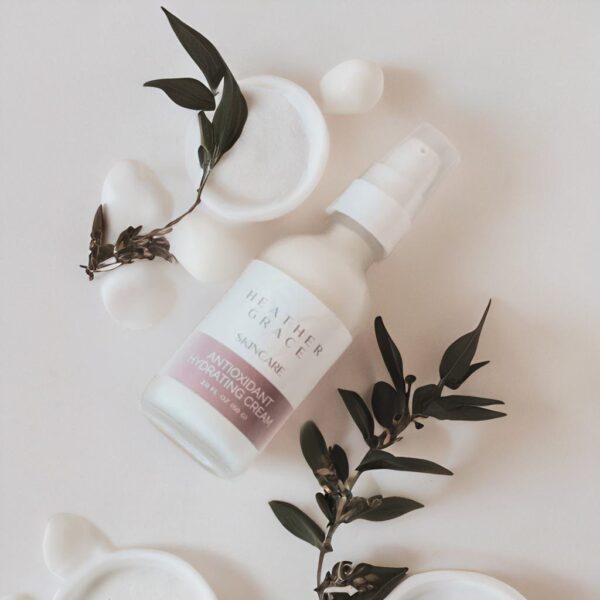 A minimalist arrangement featuring Heather Grace Skincare's Antioxidant Hydrating Cream in a white spray bottle, artistically placed alongside green leaves and white ceramic stones on a pale surface, evoking a serene and pure aesthetic.
