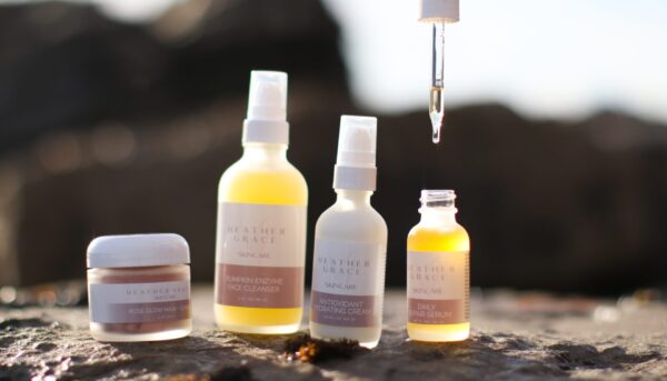A collection of Heather Grace Skincare facial products displayed on a rock with a blurred beach background. From left to right: a small white jar labeled 'ROSE GLOW MASK,' a yellow liquid in a clear spray bottle titled 'PUMPKIN ENZYME FACE CLEANSER,' a white spray bottle named 'ANTIOXIDANT HYDRATING CREAM,' and a small clear dropper bottle with golden-yellow serum identified as 'DAILY REPAIR SERUM.' A hand is holding the serum dropper above the bottle, capturing a single drop mid-air, highlighting the product's texture and consistency.