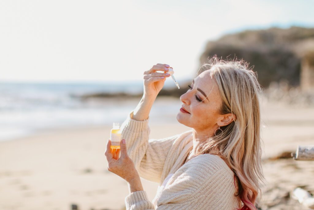 Heather Grace is holding a bottle of Daily Repair Serum up to the light, closely examining the contents. She's outdoors on a sunny beach, wearing a cream knitted cardigan over a white dress. Her expression is one of serene contentment, with her eyes gently closed and a soft smile, as she's surrounded by a tranquil coastal landscape.