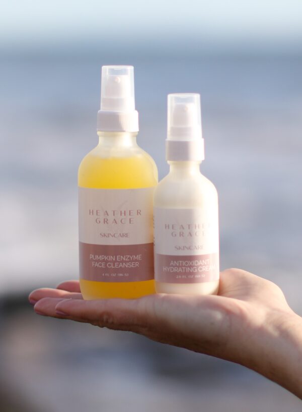 An outstretched hand holds two Heather Grace Skincare products against a serene seaside backdrop. On the left is a bottle of 'PUMPKIN ENZYME FACE CLEANSER' with a vibrant yellow liquid, and on the right, a bottle of 'ANTIOXIDANT HYDRATING CREAM.' Both bottles have clean, elegant labels and white spray nozzles. The gentle waves and soft focus on the ocean horizon evoke a sense of calm and natural beauty, reflecting the essence of the skincare line.